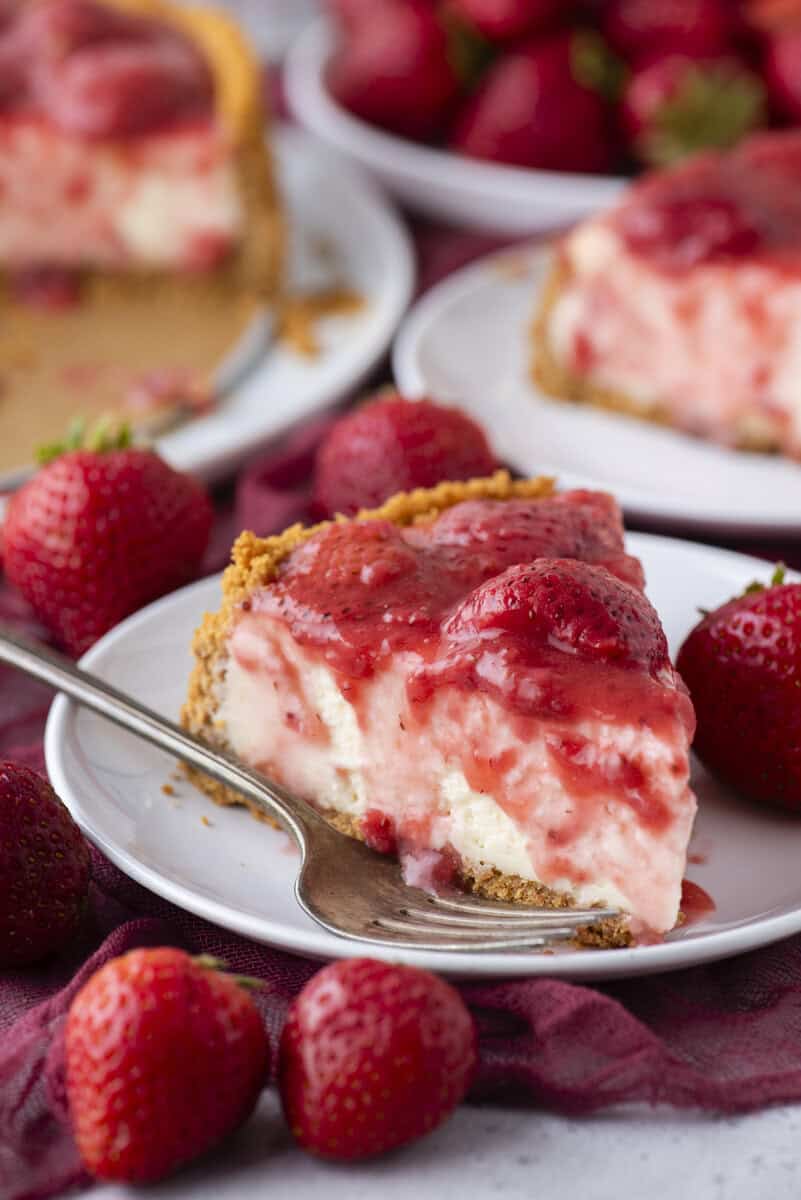A slice of strawberry cheesecake with a graham cracker crust sits on a white plate, topped with strawberry sauce and whole strawberries, surrounded by fresh strawberries and a fork. More cheesecake and strawberries are in the background.