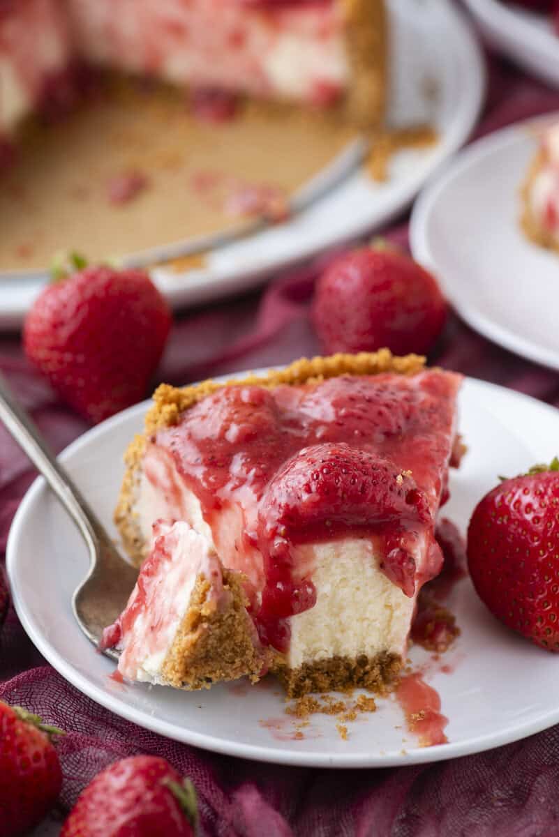 A slice of cheesecake topped with strawberry sauce and fresh strawberries on a white plate with a fork, surrounded by whole strawberries and a partially visible cheesecake in the background.