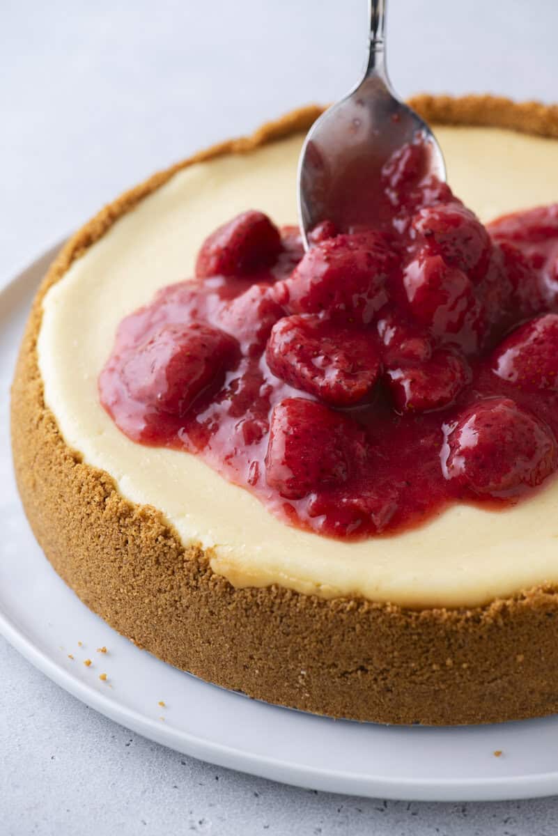 A spoon spreads glossy strawberry topping over a creamy cheesecake with a thick graham cracker crust on a white plate.