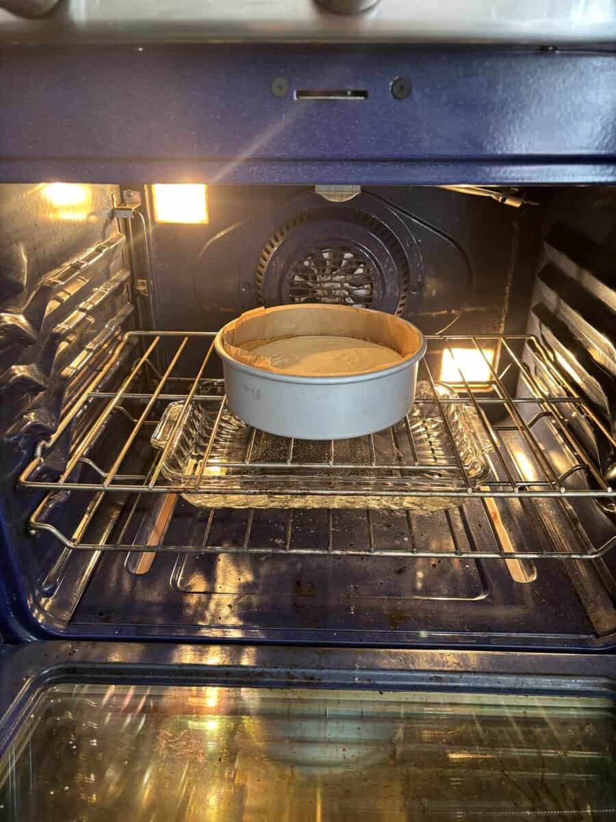 A round cake pan with batter is baking on the middle rack inside a lit oven, with the oven door open and metal racks visible.