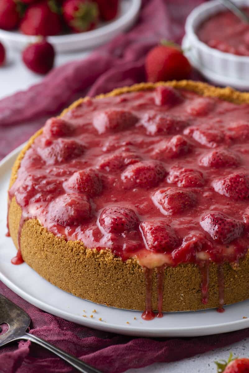 A cheesecake with a graham cracker crust, topped with a glossy strawberry sauce and whole strawberries, sits on a white plate. Some sauce is dripping down the sides. Fresh strawberries and a bowl of sauce are in the background.