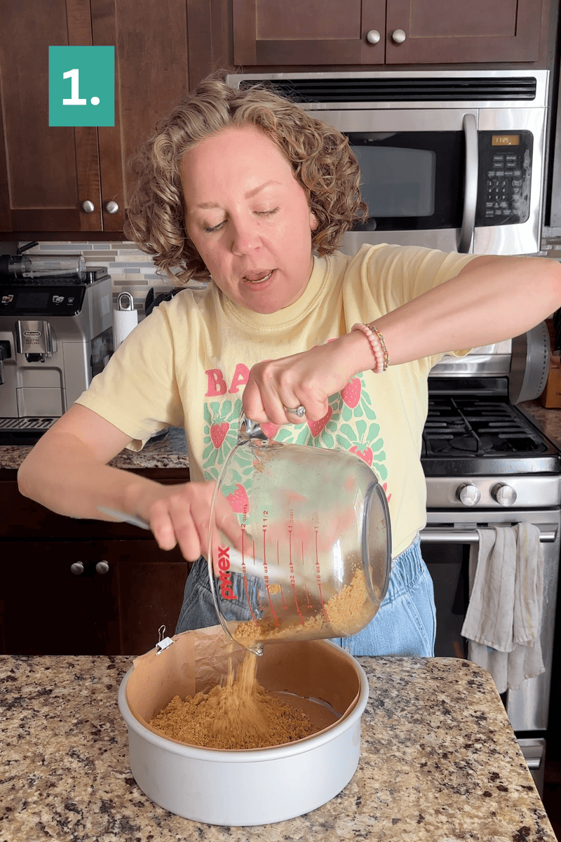 A woman pours crushed graham crackers from a glass measuring cup into a round baking pan on a kitchen counter. She stands in a modern kitchen, wearing a yellow t-shirt with a strawberry design.