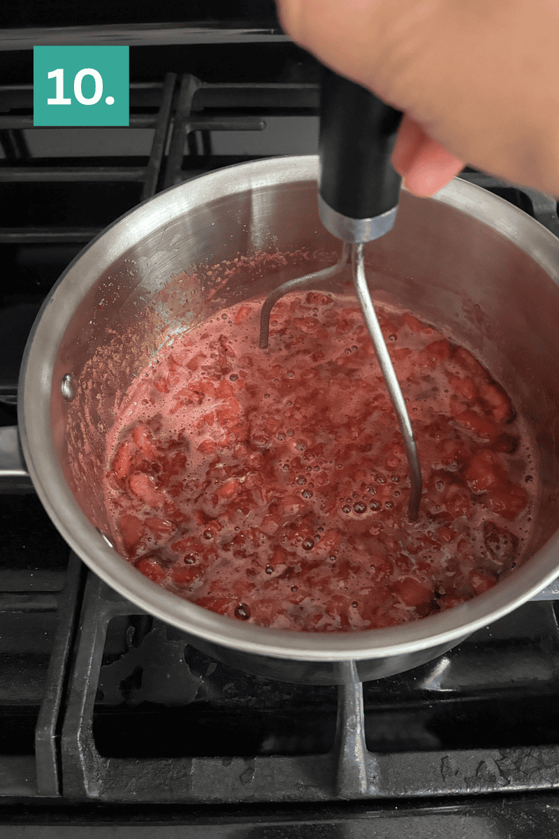 A hand uses a potato masher to mash cooked strawberries in a pot on a stove. A green box with the number 10 is in the top left corner.