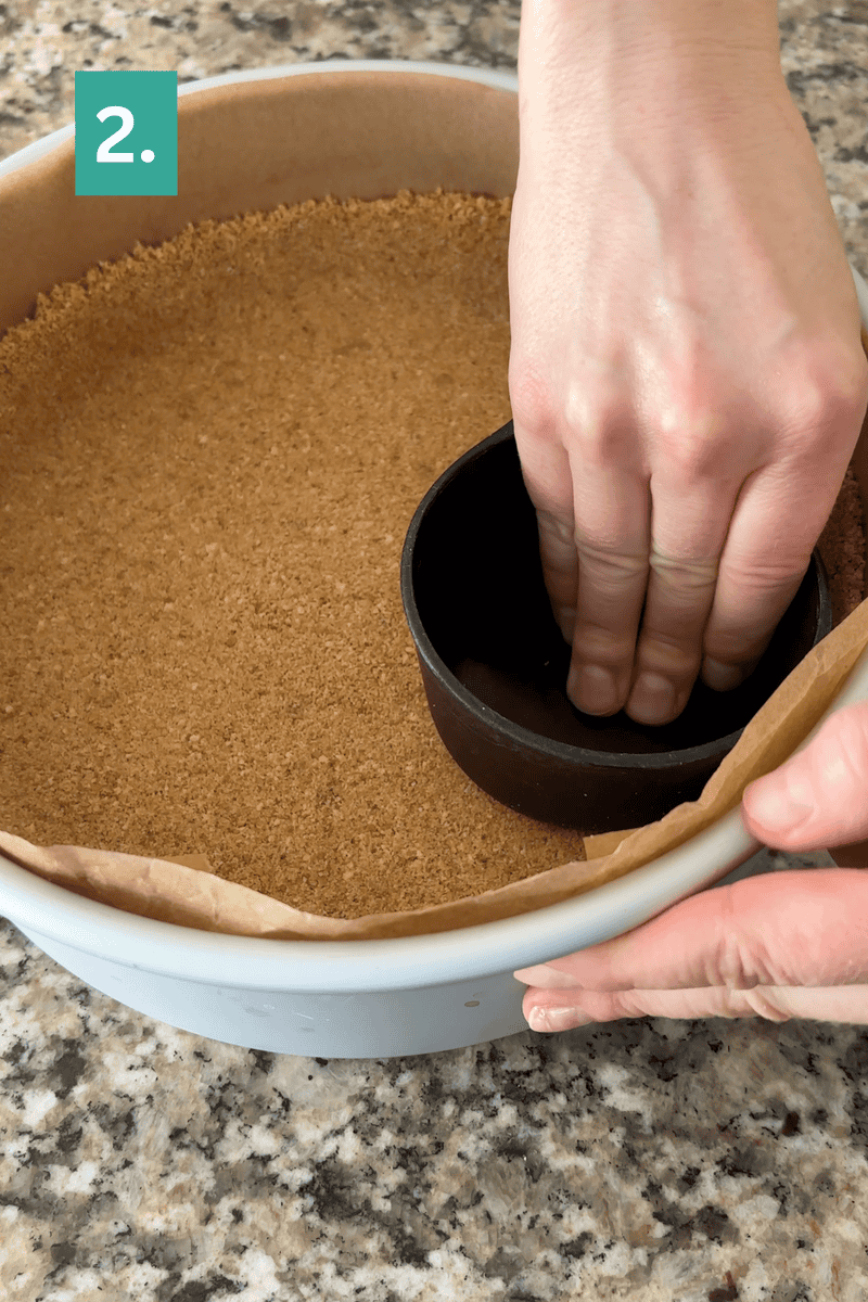 A person presses a measuring cup into a pie pan lined with parchment and filled with a crumb crust, shaping it evenly. The countertop is granite, and a green square with the number 2 is in the top left corner.