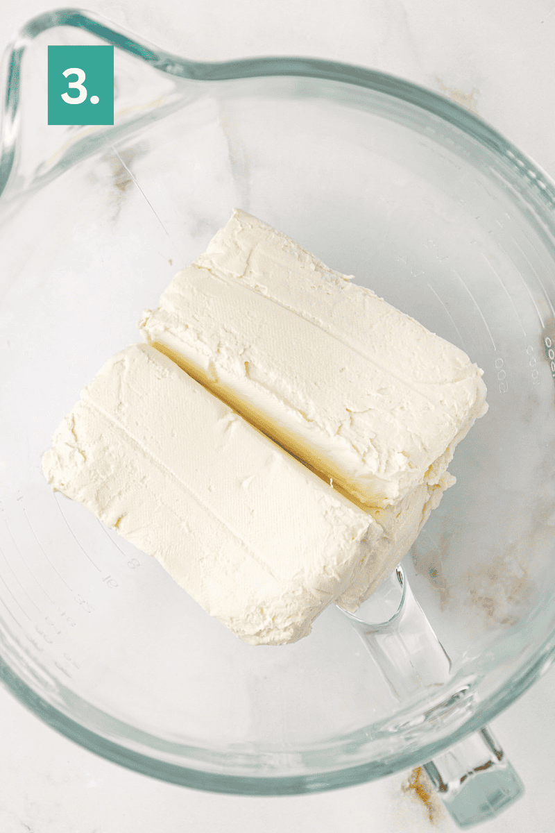 Two blocks of cream cheese in a clear glass mixing bowl, viewed from above, with a green box labeled “3.” in the top left corner.