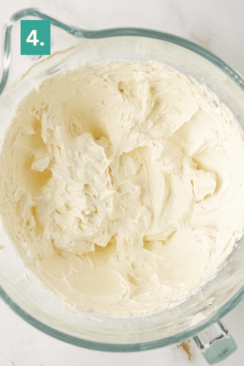 A large glass mixing bowl filled with creamy, whipped buttercream frosting. A green box in the top left corner displays the number 4, indicating a step in a recipe.