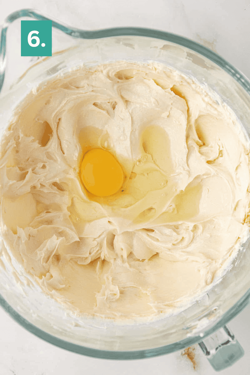 A glass mixing bowl filled with creamy, pale batter and a raw egg cracked on top. A green square with the number 6 is in the upper left corner of the image.