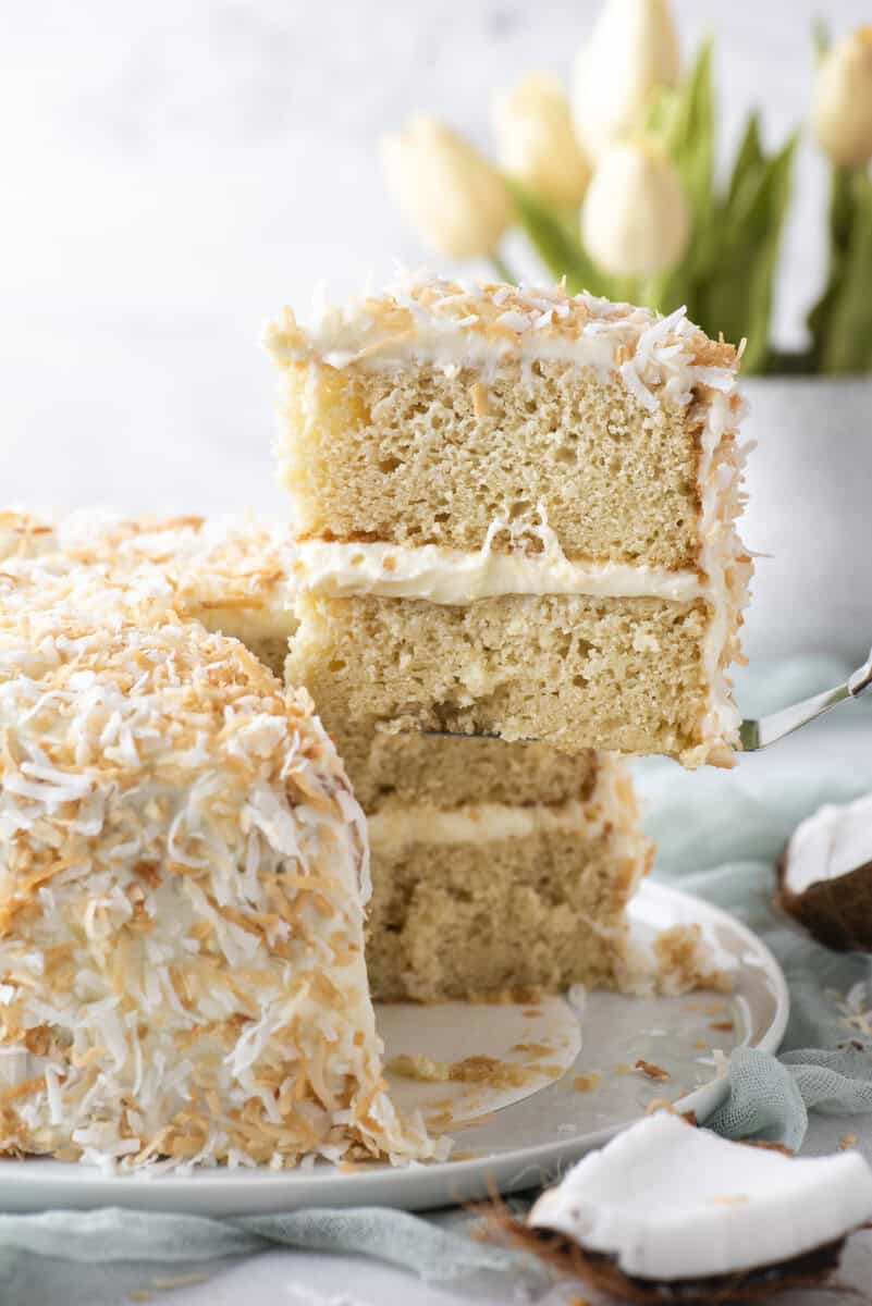 A slice of coconut layer cake with cream filling and toasted coconut flakes is being lifted from the whole cake. White tulips and a coconut shell are in the background.