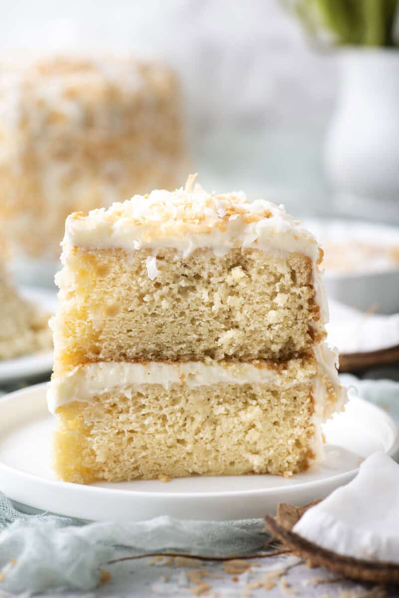 A close-up of two stacked slices of coconut cake with white frosting and toasted coconut flakes on top, served on a white plate.