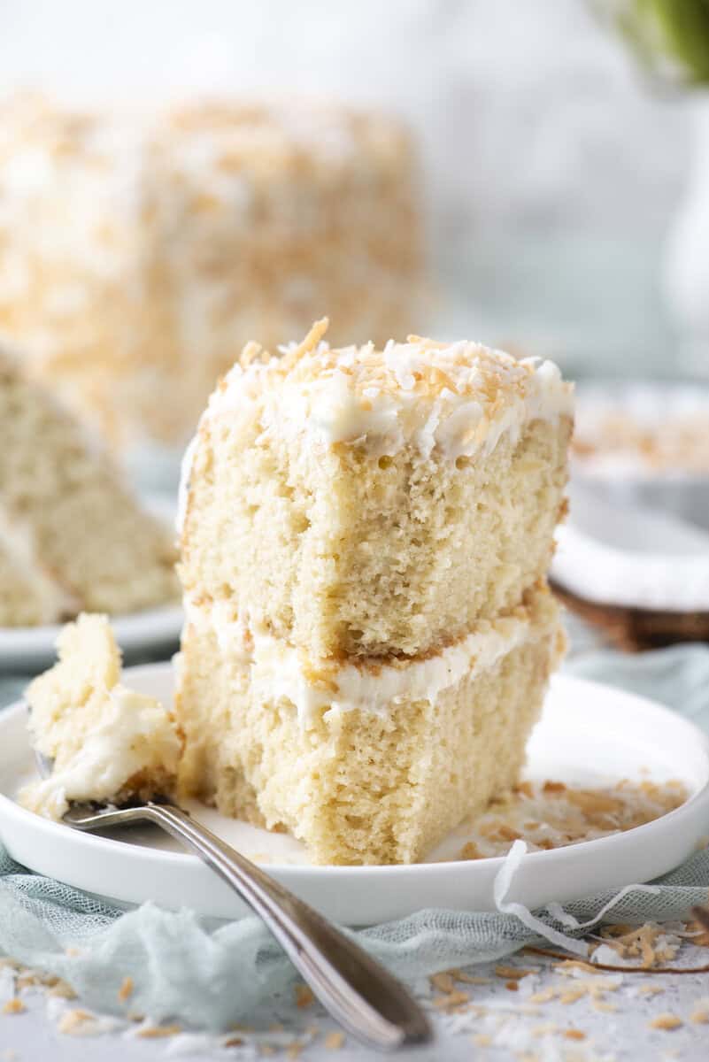 Two slices of coconut cake with white frosting are stacked on a white plate, topped with toasted coconut. A fork with a bite of cake rests beside the slices. The background includes more cake and a soft turquoise cloth.
