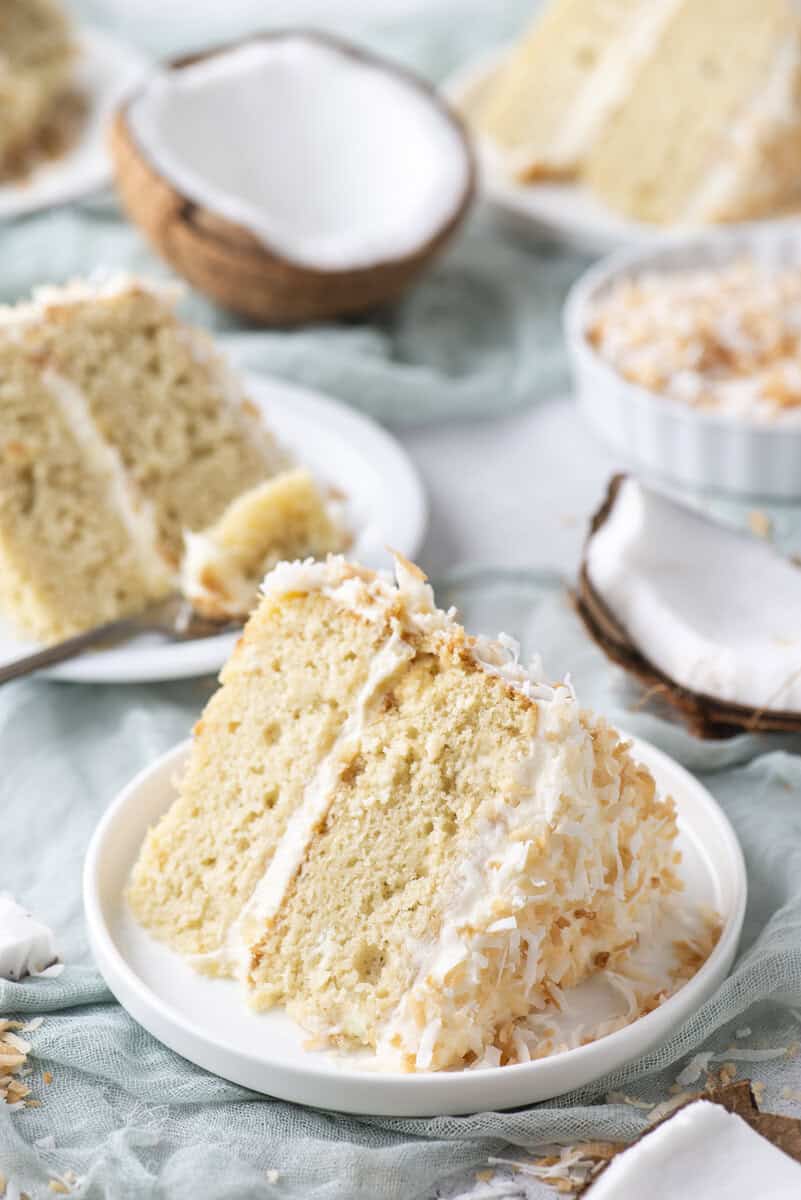 A slice of coconut cake with white frosting and shredded coconut on a plate, surrounded by coconut halves and more cake slices in the background.