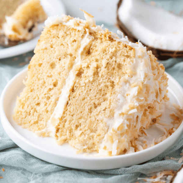 A slice of coconut cake with white frosting and toasted coconut flakes sits on a white plate, with a coconut half and more cake visible in the background.