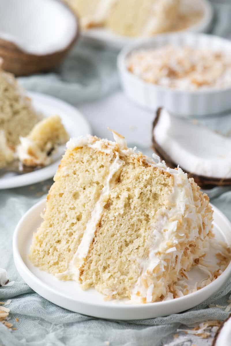 A slice of coconut layer cake with white frosting and shredded coconut sits on a white plate, with more cake and coconut pieces in the background.
