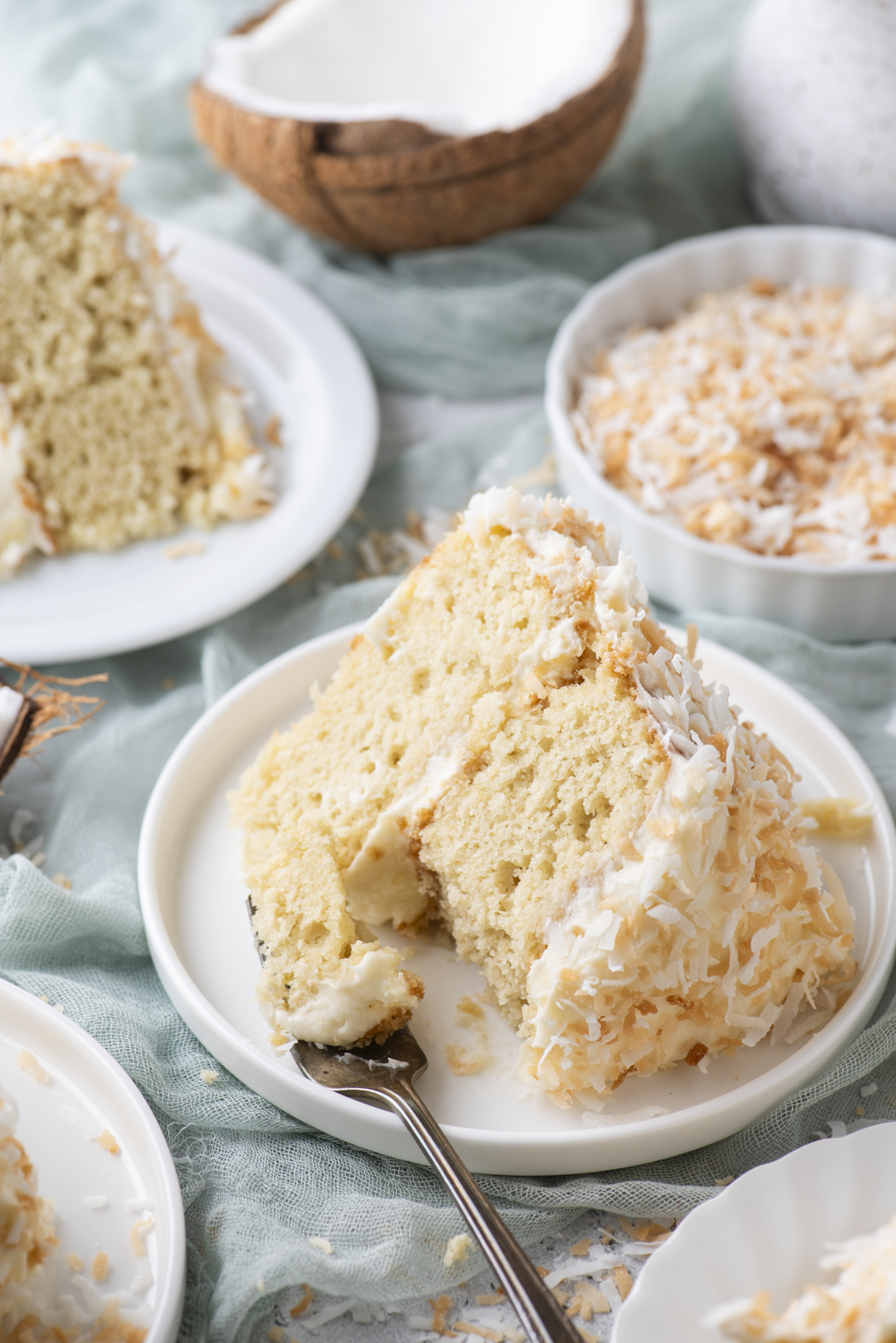 A slice of coconut cake with creamy frosting and toasted coconut flakes sits on a white plate, with a fork beside it. In the background are more cake slices, a bowl of toasted coconut, and a halved coconut.