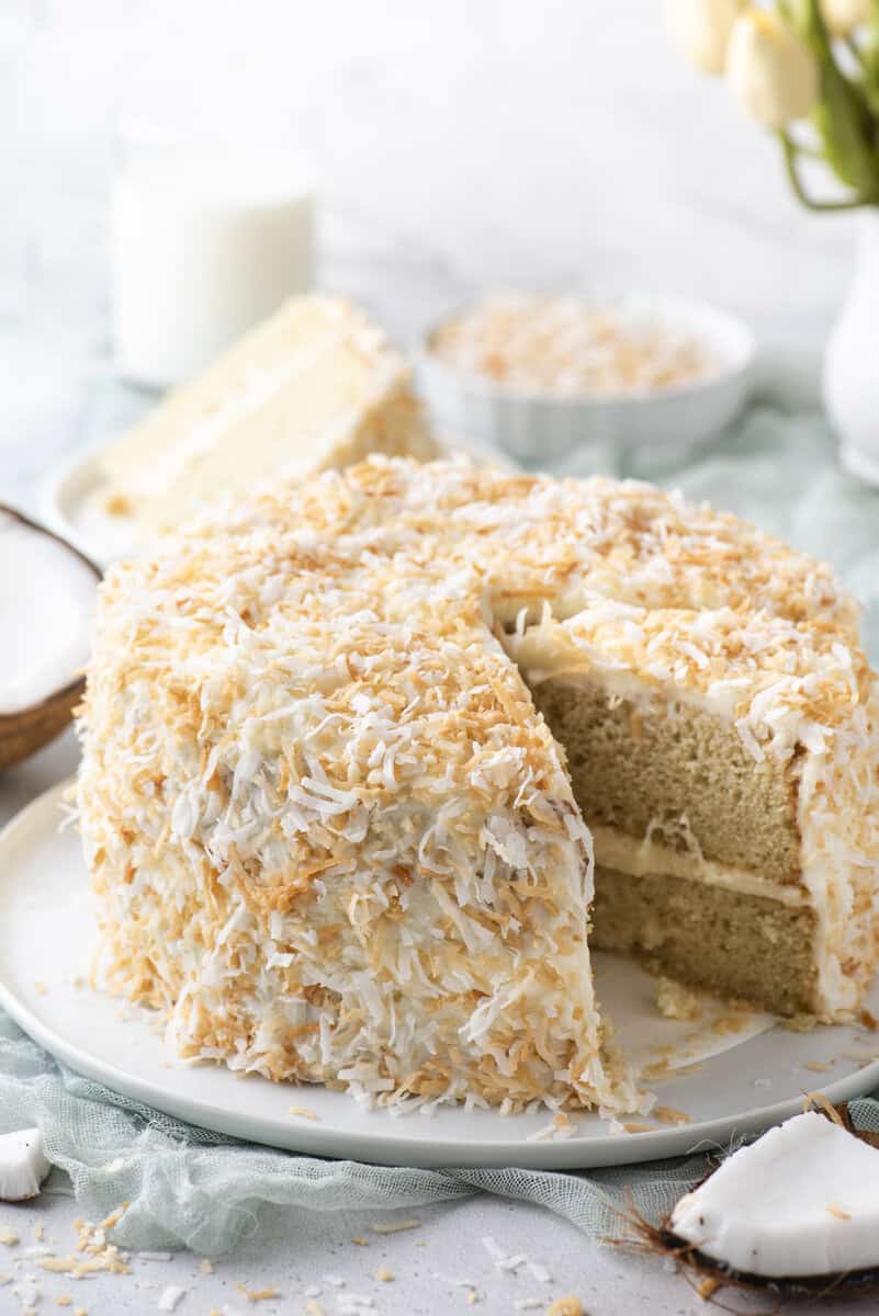 A round layer cake covered in toasted coconut flakes sits on a white plate, with one slice cut and slightly pulled out. In the background are a glass of milk, a bowl of coconut, and a vase of flowers.