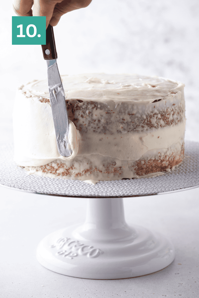 A hand uses an offset spatula to spread frosting on a partially frosted cake set on a white cake stand. The number 10. appears in a green box in the top left corner.