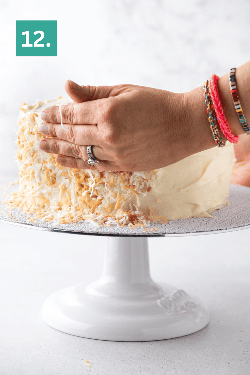A hand presses toasted coconut flakes onto the side of a frosted cake on a white stand. A green box with “12.” is in the top left. The person wears colorful beaded bracelets.