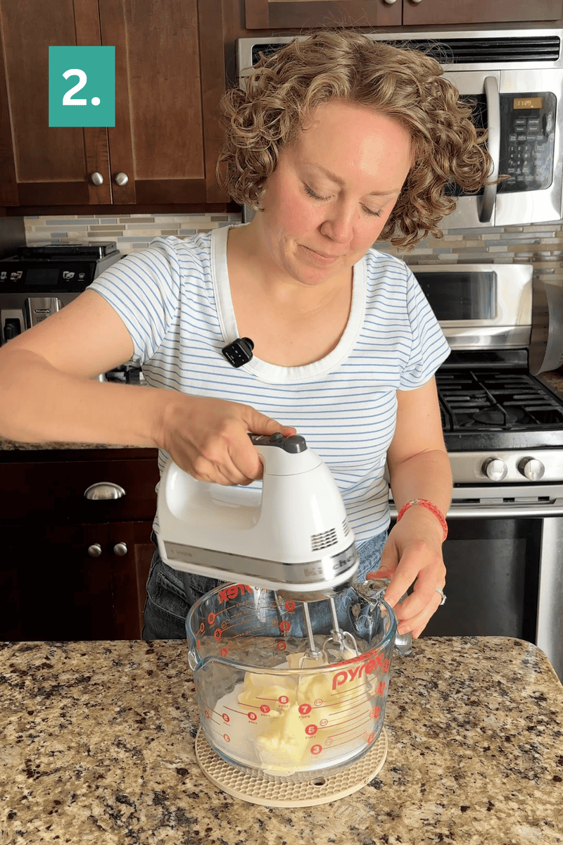 A woman uses a hand mixer to blend ingredients in a large glass measuring cup on a kitchen counter. A green box with the number 2 is in the upper left corner.