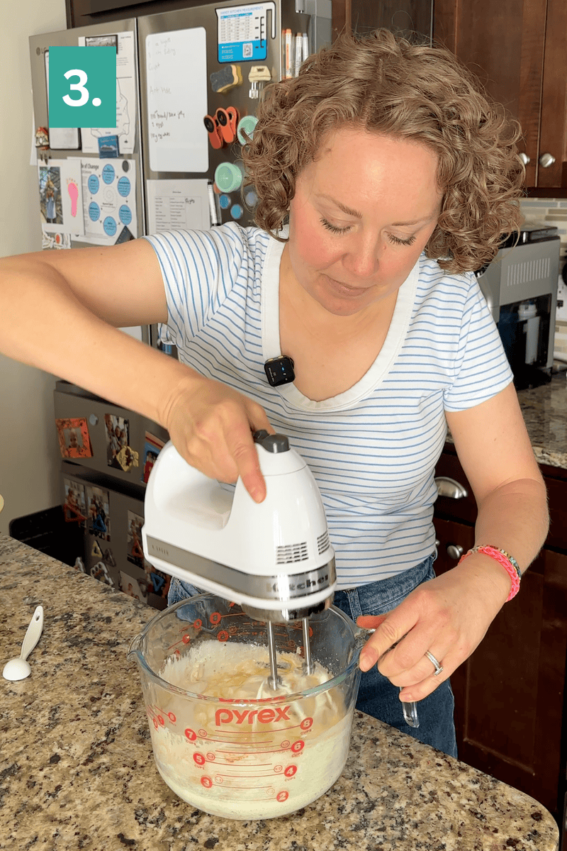 A woman mixes ingredients with a hand mixer in a large Pyrex measuring cup on a kitchen counter. She wears a striped shirt and stands near a fridge covered in photos. A green 3. label is in the top left corner.