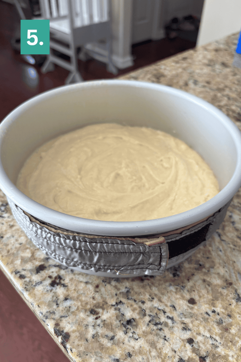 A round cake pan with light batter inside sits on a granite countertop. The pan is wrapped with a damp cake strip. A green box in the corner displays the number 5.