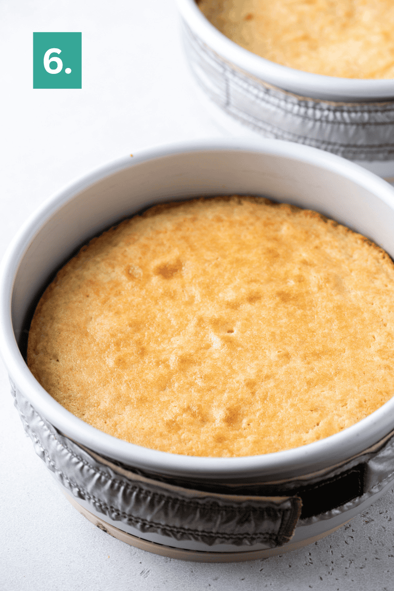 Baked cake in a round white pan with a golden top, wrapped in a gray oven mitt, sitting on a white surface. A green box with the number 6 is in the corner.