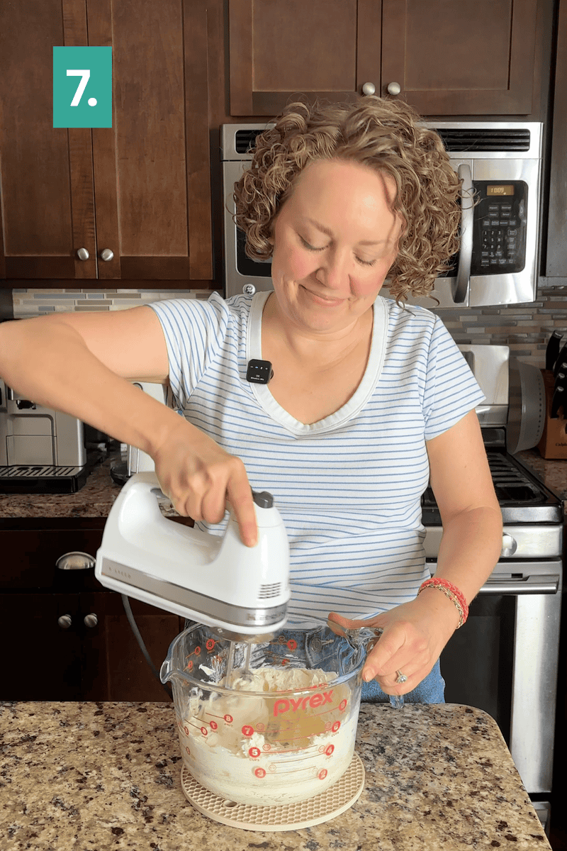 A person with curly hair uses a hand mixer to blend ingredients in a Pyrex bowl on a kitchen counter. A green box with the number 7 is in the upper left corner.