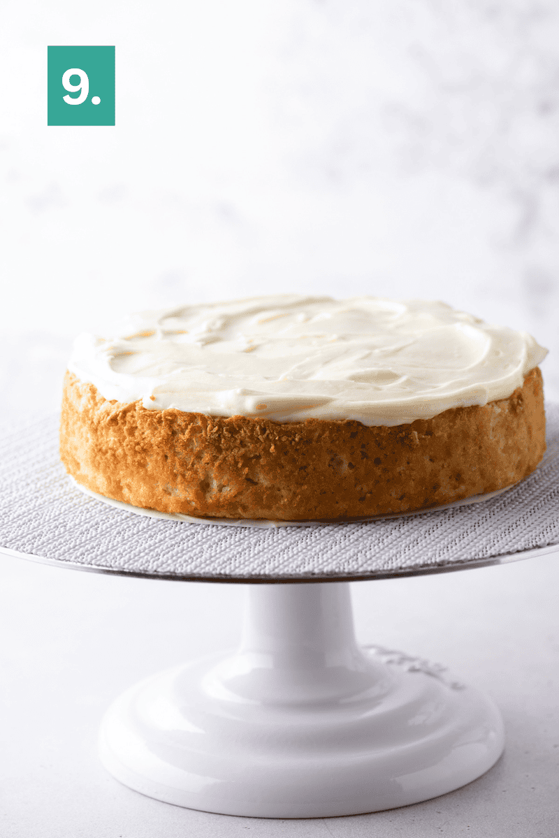 A single-layer cake with white frosting sits on a white cake stand, with a green box displaying the number 9 in the top left corner.