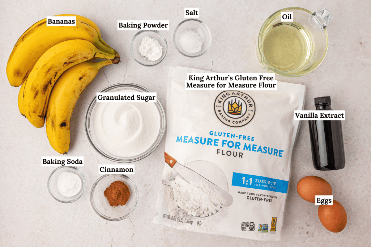 A flat lay of banana bread ingredients: bananas, King Arthur’s gluten free flour, sugar, eggs, oil, vanilla, cinnamon, baking soda, baking powder, and salt, each in bowls or containers on a white surface.