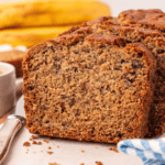 A close-up of sliced banana bread on a white surface, with bananas and a knife in the background. The bread looks moist, dense, and has a golden-brown crust.
