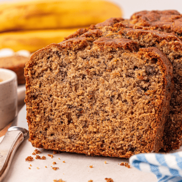 A close-up of sliced banana bread on a white surface, with bananas and a knife in the background. The bread looks moist, dense, and has a golden-brown crust.