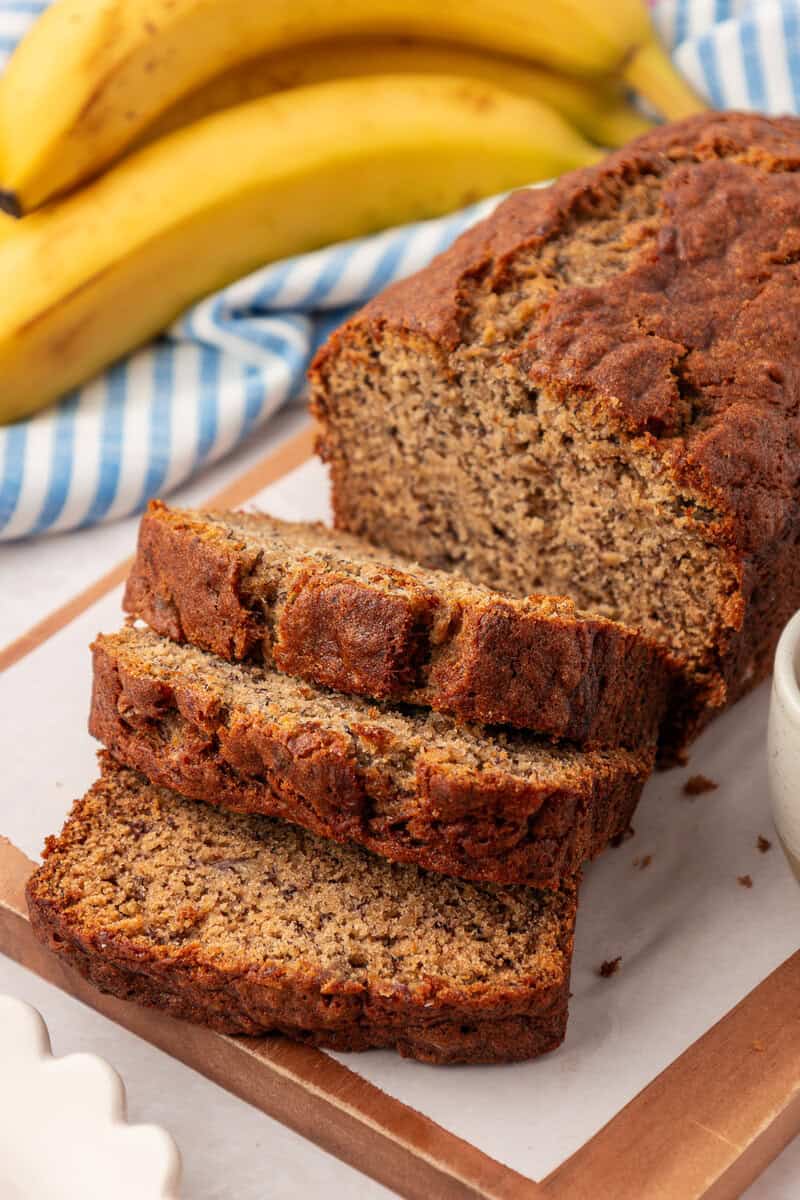 A sliced loaf of banana bread sits on a wooden cutting board, with fresh bananas and a blue-striped cloth in the background.