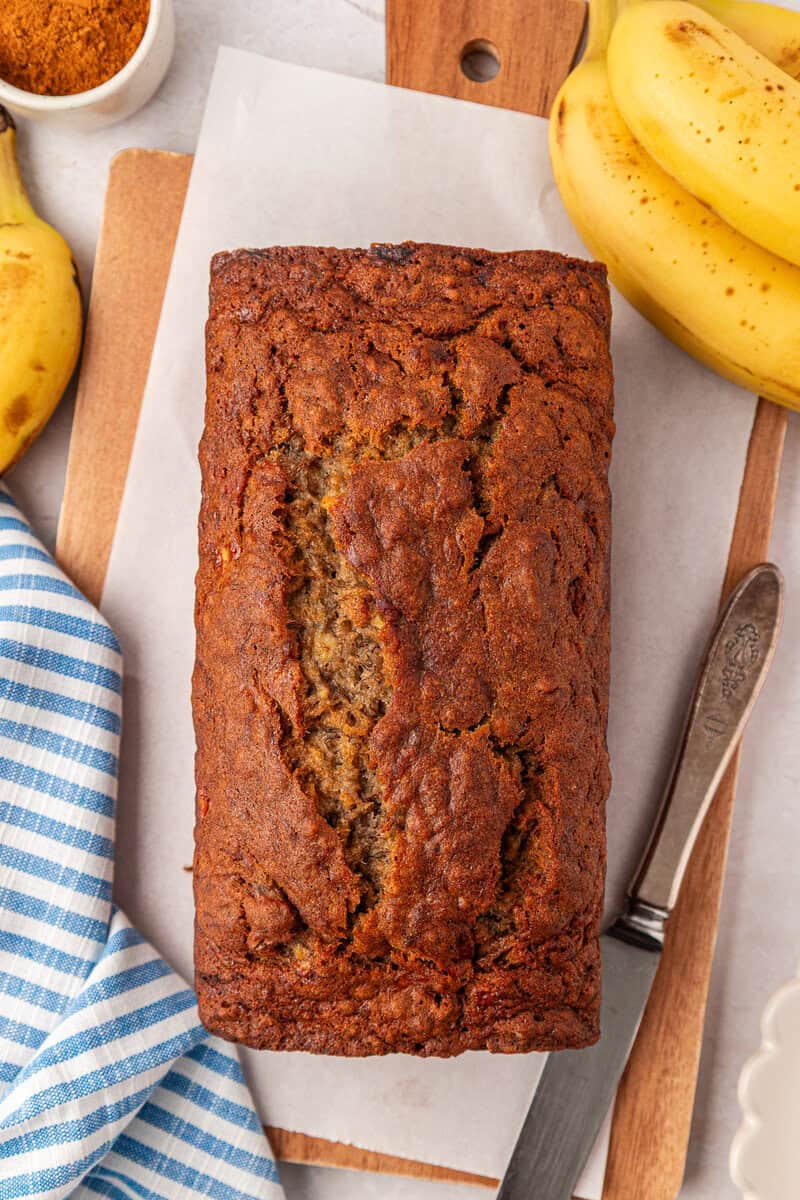 A loaf of banana bread sits on parchment paper, surrounded by bananas, a knife, a wooden board, and a blue-striped cloth.