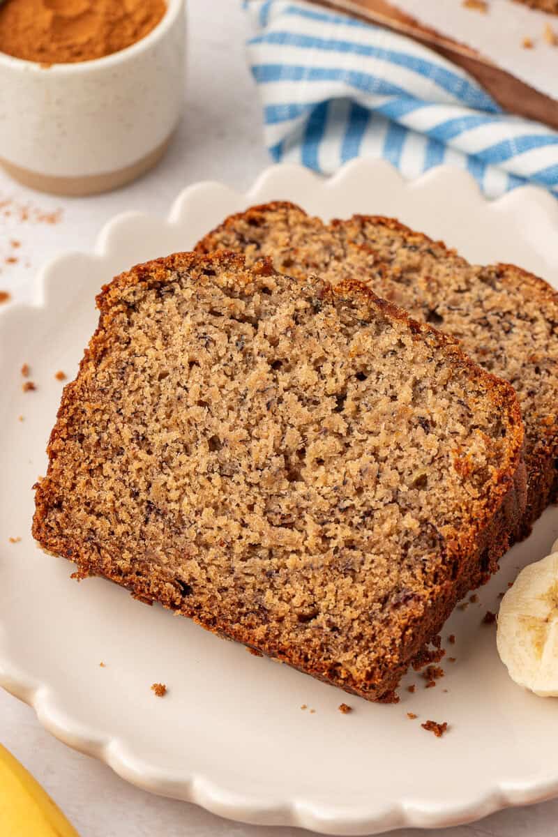 Two slices of banana bread sit on a white scalloped plate, with a few banana slices nearby. A small bowl of cinnamon and a blue striped cloth are in the background.