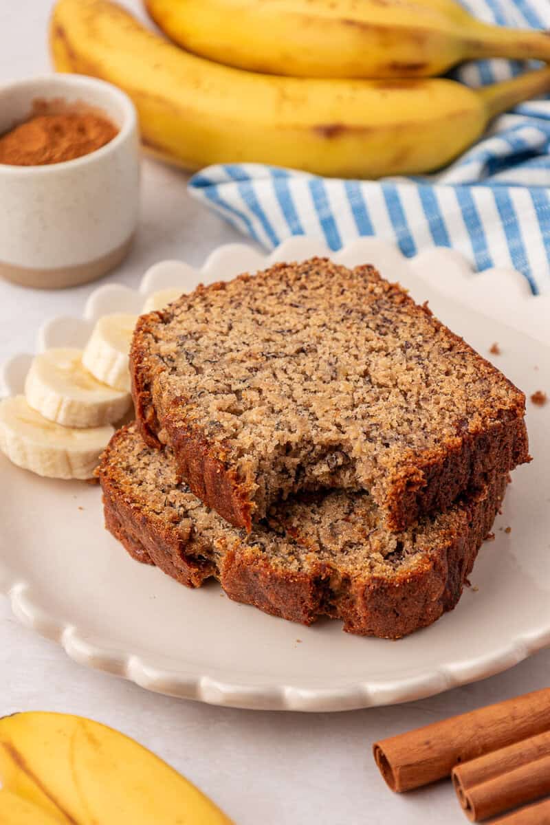 Two slices of banana bread on a plate with banana slices, cinnamon sticks, whole bananas, and a bowl of cinnamon in the background.