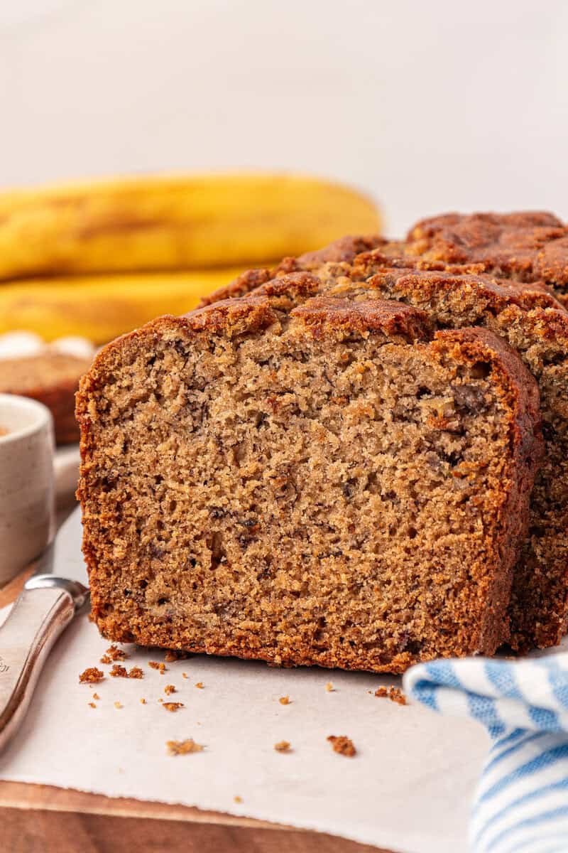 A close-up of sliced banana bread on parchment paper, with ripe bananas and a cup in the background. A blue and white cloth is partially visible in the foreground.