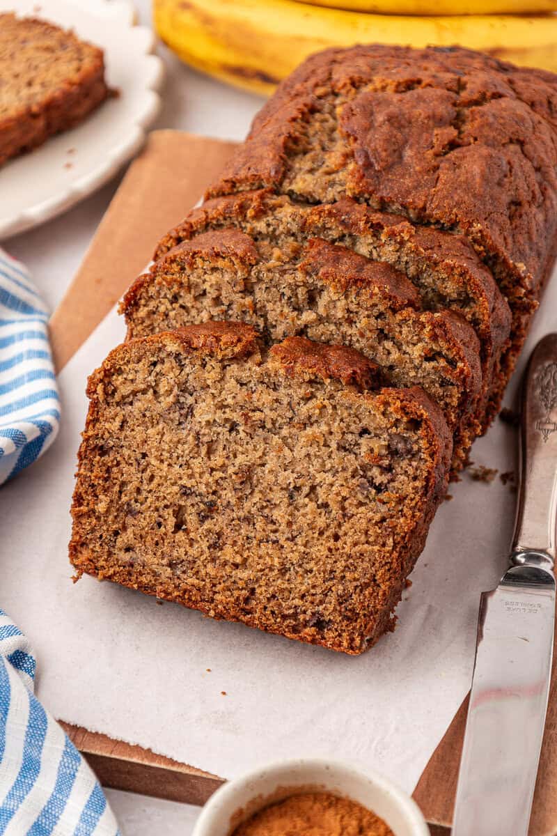 A loaf of banana bread sliced on a cutting board, with a knife beside it and a bunch of bananas in the background. A blue-striped towel and a small bowl of cinnamon are also visible.