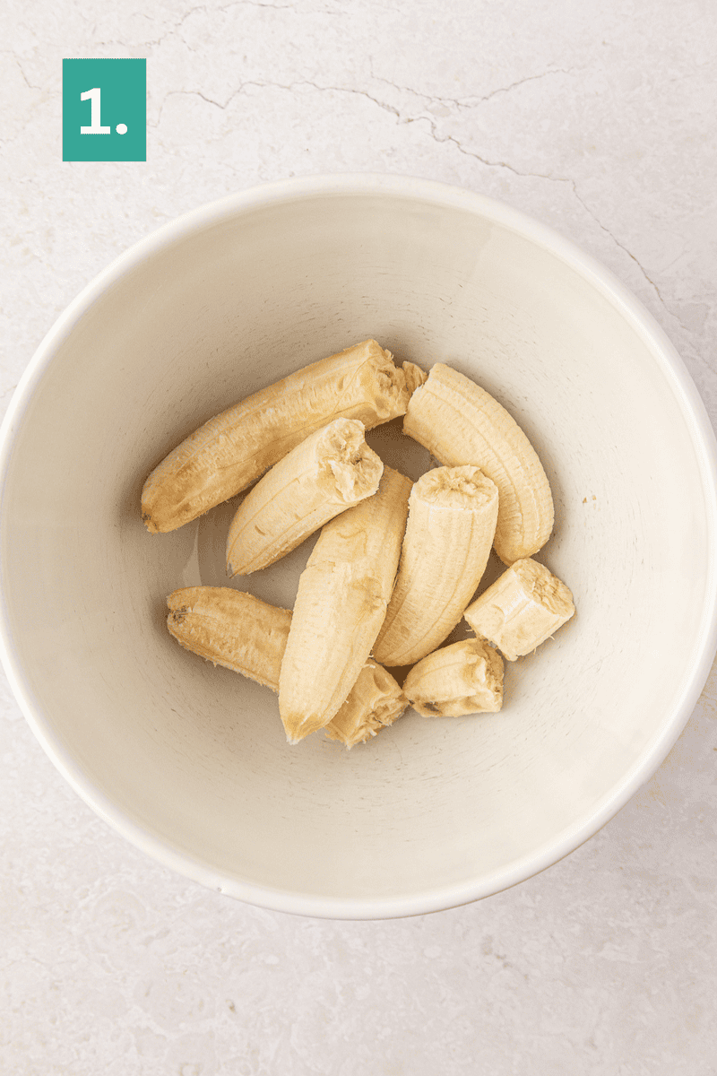 A white bowl containing peeled and halved bananas sits on a light-colored surface. A green box with the number 1 is in the top left corner.