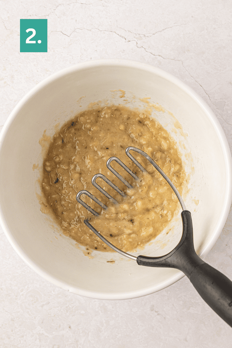 A bowl of mashed bananas with a potato masher inside. A green box with the number 2 is in the top left corner of the image.