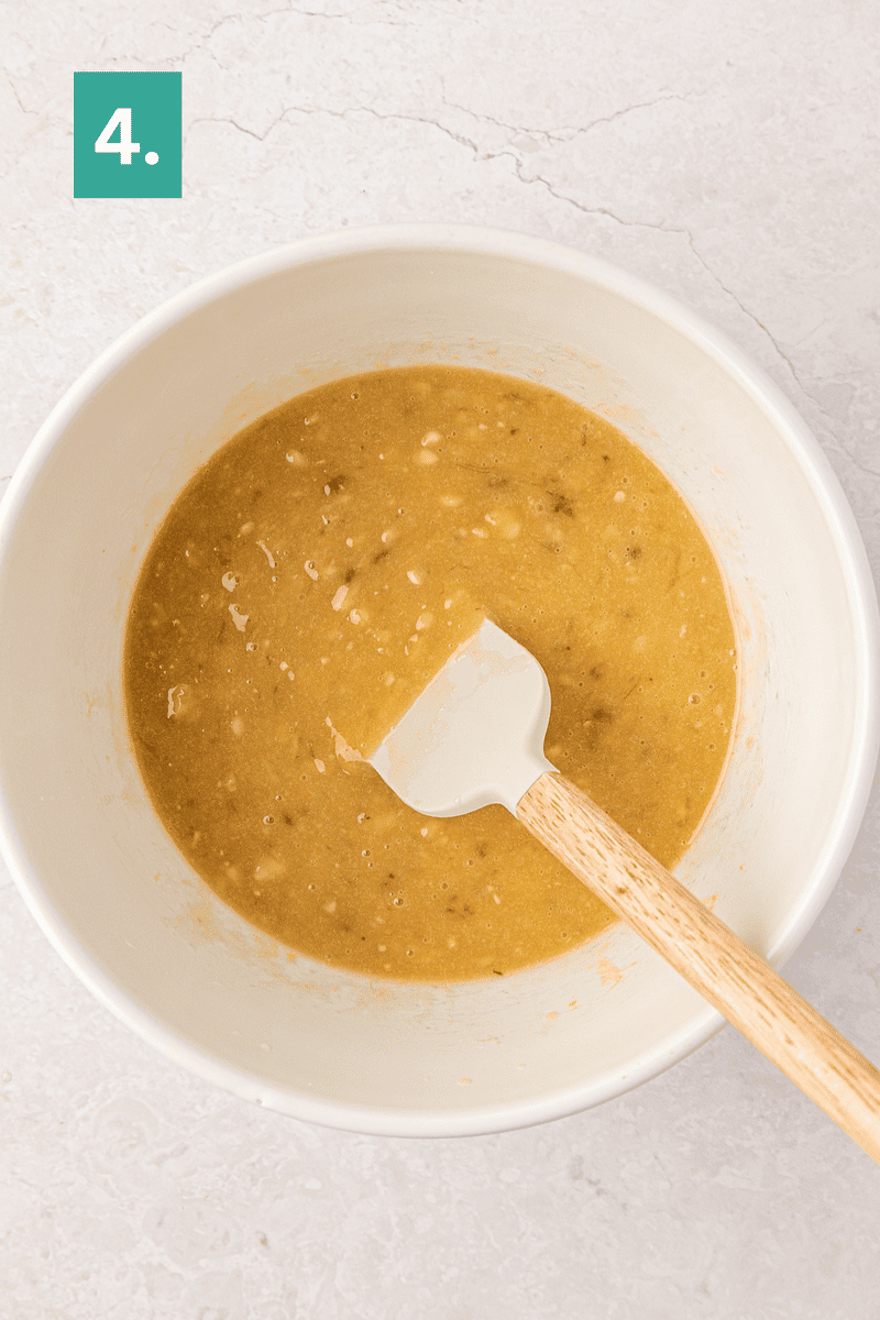 A white mixing bowl with a banana bread batter and a spatula inside. A green box with the number 4 is in the top left corner. The bowl sits on a light countertop.