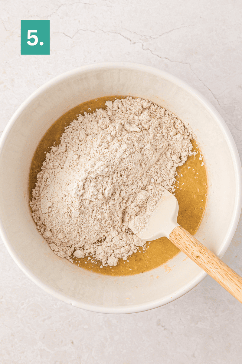 A mixing bowl with flour sitting on top of a wet batter, being stirred with a white spatula. A green box with the number 5 is in the top left corner.