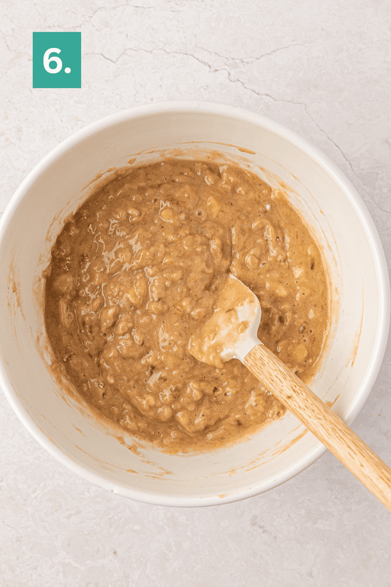 A white bowl with banana bread batter and a spatula inside. A green box with the number 6 is in the top left corner. The bowl sits on a light textured surface.