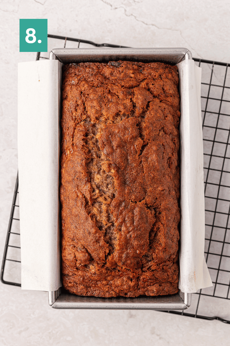 A loaf of banana bread in a parchment-lined metal pan sits on a cooling rack. A green square with the number 8 is in the top left corner of the image.
