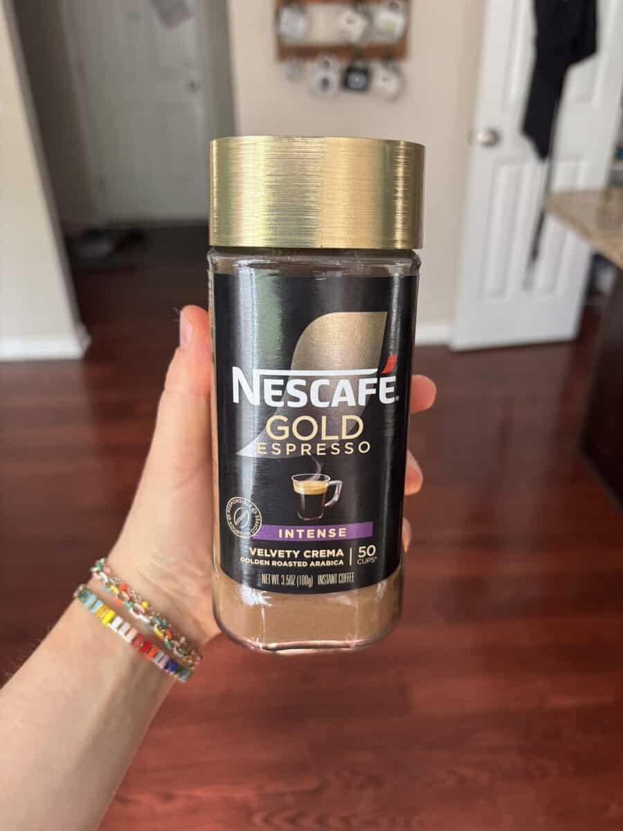 A hand holds a jar of Nescafé Gold Espresso Intense coffee. The jar has a gold lid and a black label with a coffee cup image. The background shows a kitchen with wooden floors and white doors.