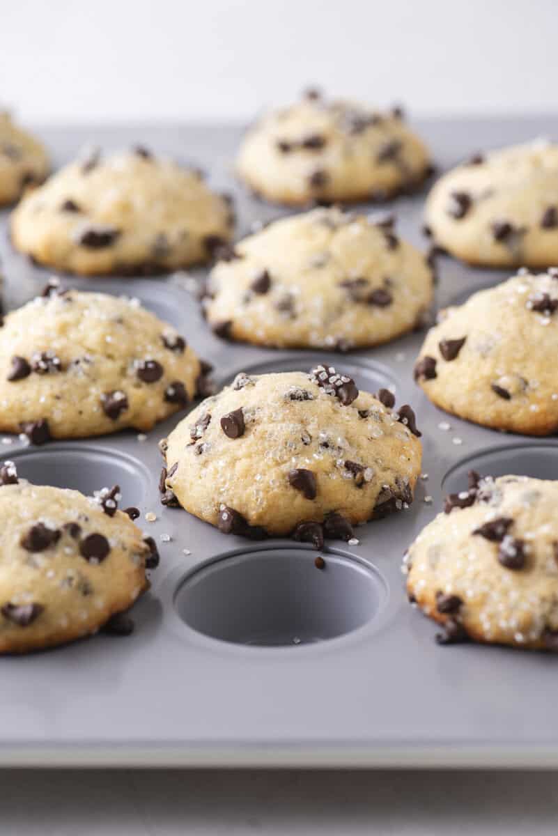 A close-up of freshly baked chocolate chip muffins in a gray muffin tin, with golden tops and sprinkled sugar, some chocolate chips scattered on the surface. One muffin slot is empty.