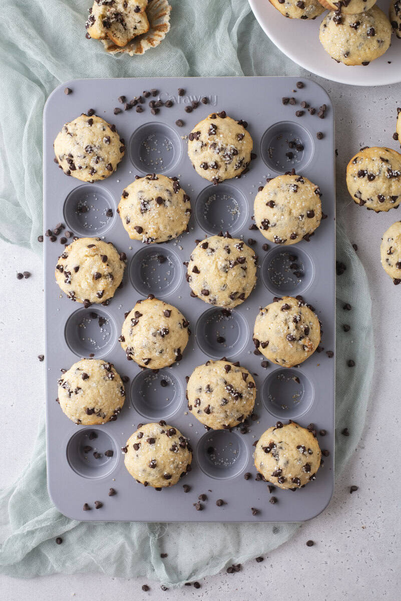 A muffin tin filled with baked mini chocolate chip muffins sits on a light blue cloth, with chocolate chips scattered around and more muffins on a white plate nearby.