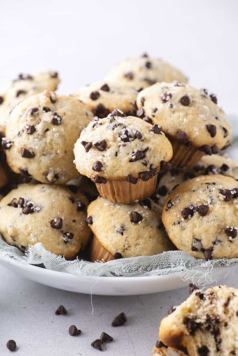 A plate piled with mini chocolate chip muffins, topped with coarse sugar and scattered chocolate chips, sits on a light fabric and white surface.