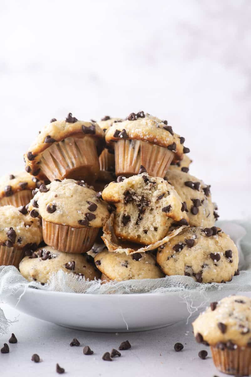 A white plate piled high with mini chocolate chip muffins, some with bites taken out, showing the soft, chocolate-filled inside. A light blue cloth is under the plate, and chocolate chips are scattered around.