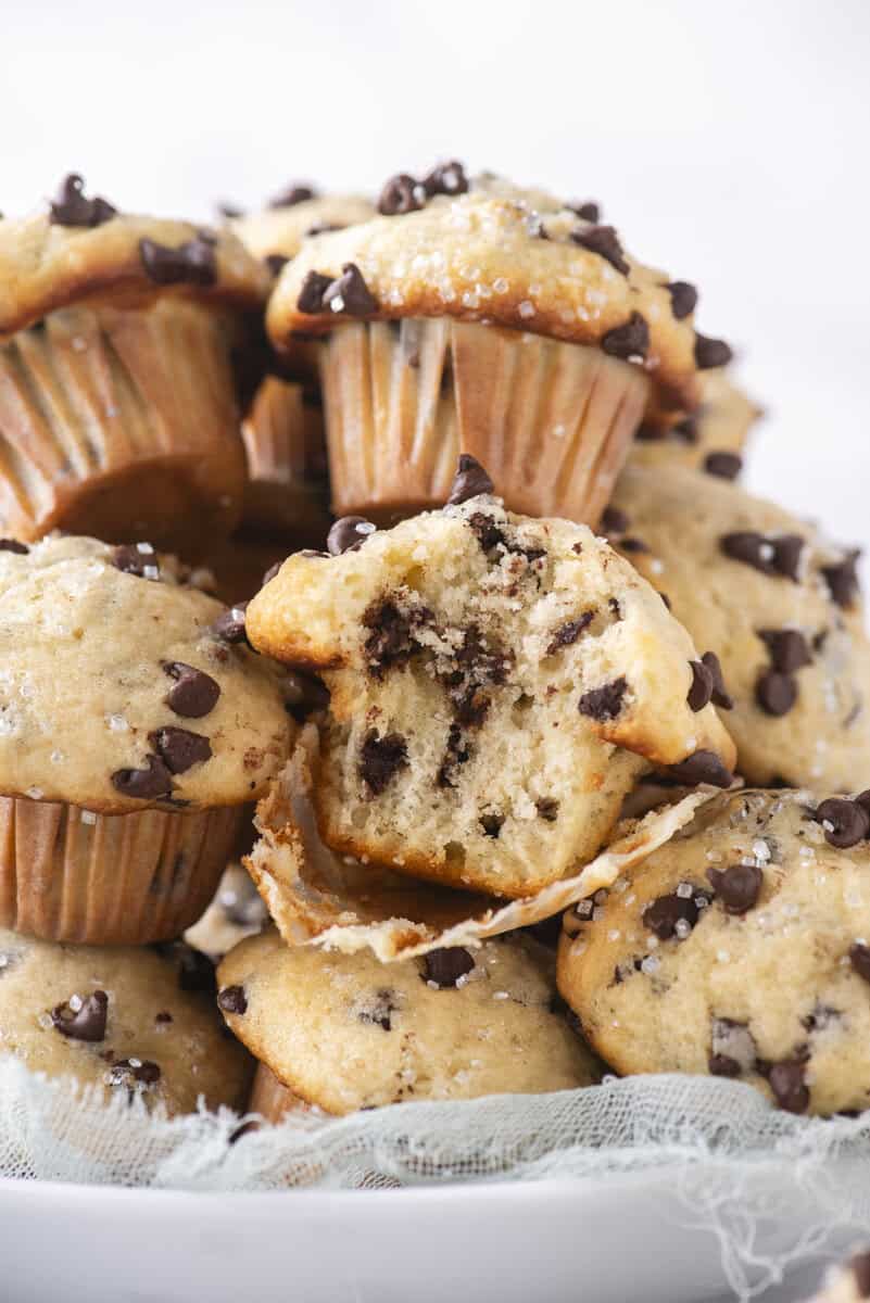 A close-up of a plate stacked with chocolate chip muffins, with one muffin in the center split open to show the soft, chocolate-filled interior. The muffins are sprinkled with sugar crystals.