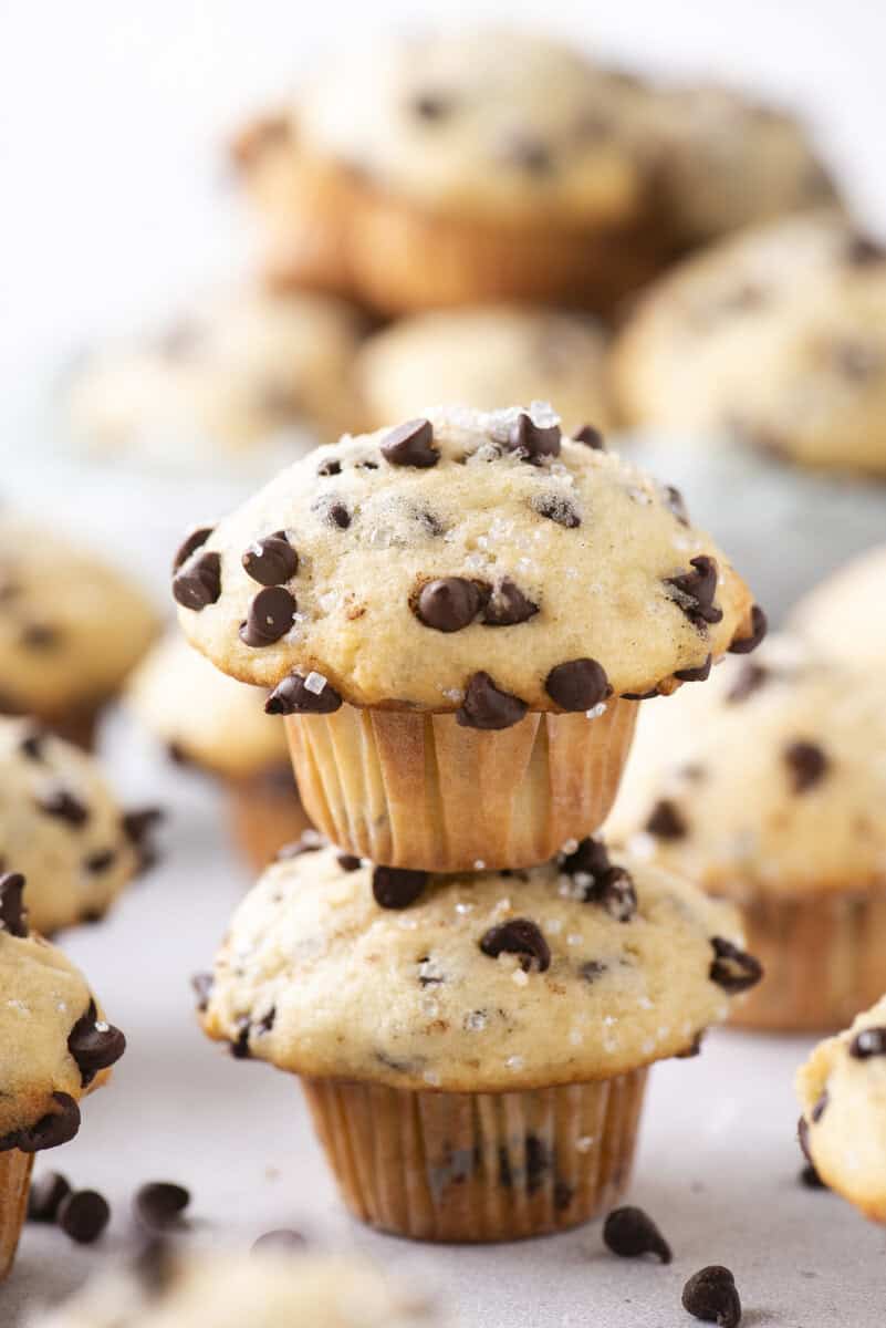 A close-up of two chocolate chip muffins stacked on top of each other, surrounded by more muffins in the background, all topped with sugar and chocolate chips.