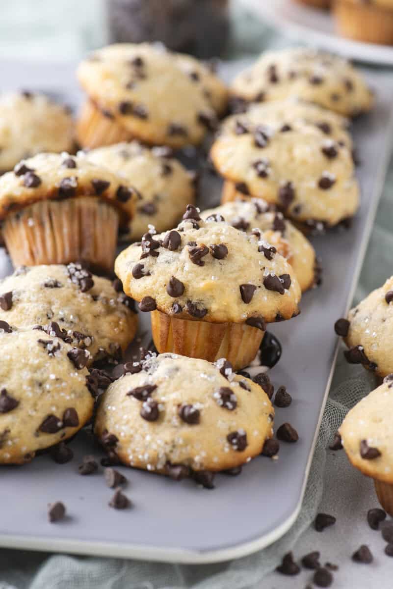 A tray of freshly baked chocolate chip muffins, topped with sparkling sugar and chocolate chips, sits on a light-colored surface with a soft cloth nearby. Some muffins are slightly overfilled and have chocolate chips scattered around them.